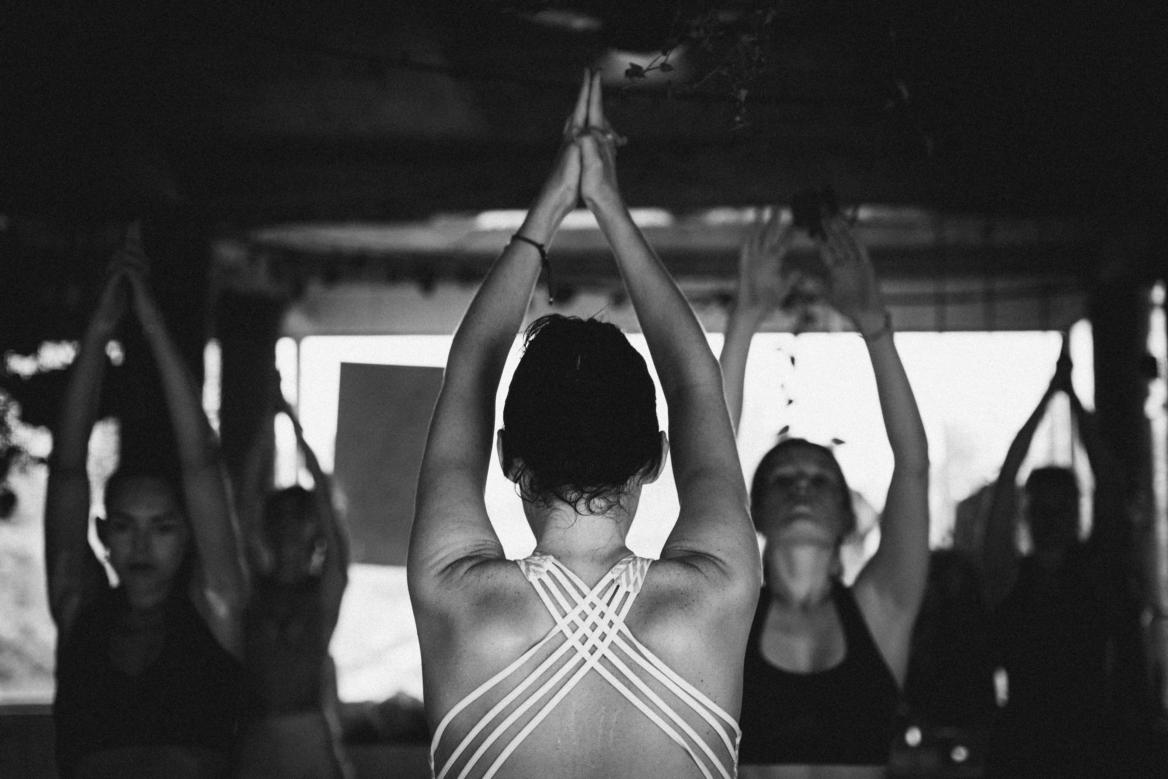 A yoga class in black and white, a woman in front raising her joined hands overhead as the group follows